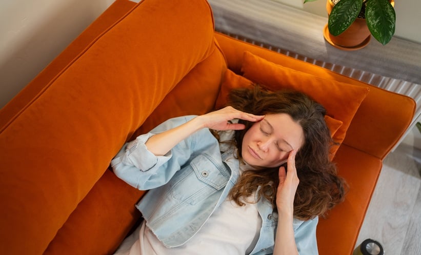 Woman resting on a sofa holding her temples, representing chronic eye pain symptoms and discomfort linked to chronic ocular surface pain.