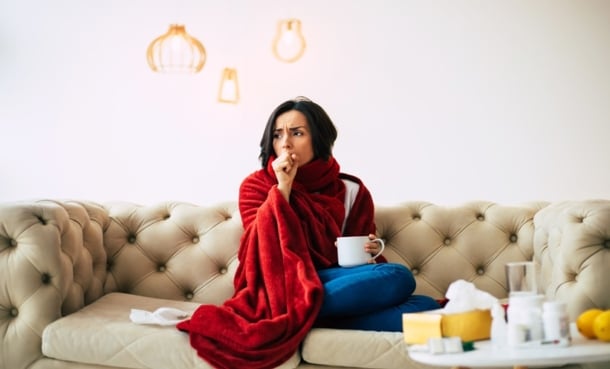 Woman wrapped in a red blanket on a sofa, holding a mug and coughing, with tissues and cold remedies nearby showing flu symptoms.