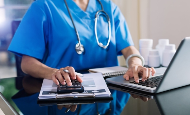 Healthcare professional in blue scrubs using a calculator and laptop to review medical costs and treatment data for a cost-effectiveness analysis.