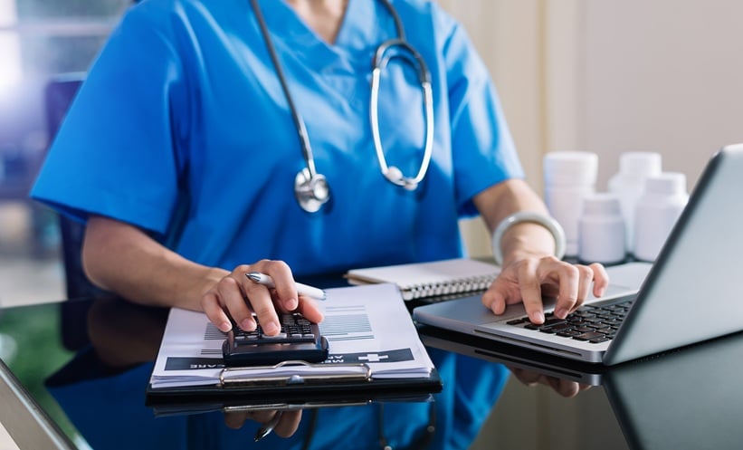 Healthcare professional in blue scrubs using a calculator and laptop to review medical costs and treatment data for a cost-effectiveness analysis.