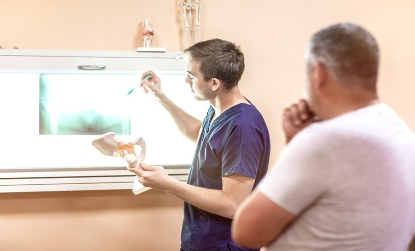 Doctor points to a hip X ray while explaining hip involvement in axial spondyloarthritis to a patient, holding a pelvis and femur anatomy model.