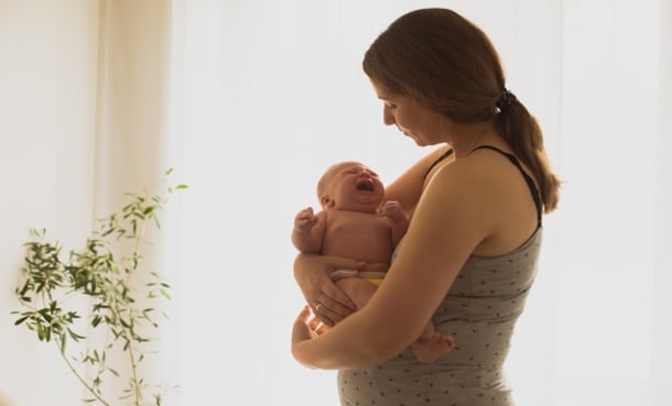 Mother holding a crying newborn baby by a bright window, representing postpartum care and follow-up after cesarean delivery.