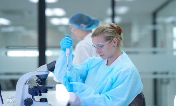 Scientist standing behind a microscope while holding up a vial and a pipette.