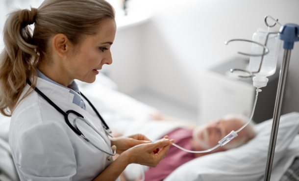 Doctor checking out an IV infusion bag in a hospital setting, while a patient lies in a hospital bed in the background.
