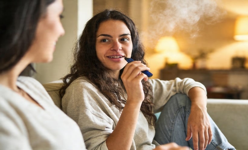 Two women seated indoors, one holding an e-cigarette while visible vapor rises, illustrating vaping and cancer risk.