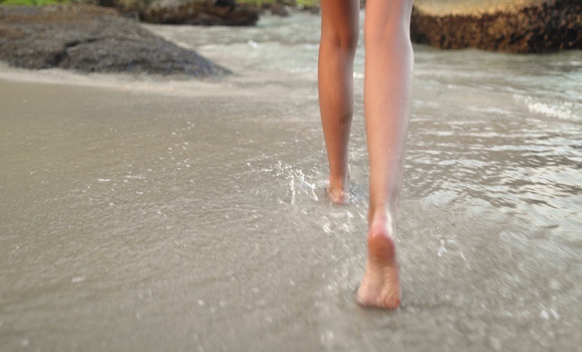 Person walking barefoot through shallow seawater along a sandy beach.