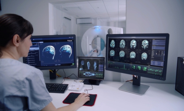 Female radiologist sits in control room and watches at monitors with displayed brain scans results. Professional doctor controls MRI or CT or PET scan with patient undergoing procedure at background