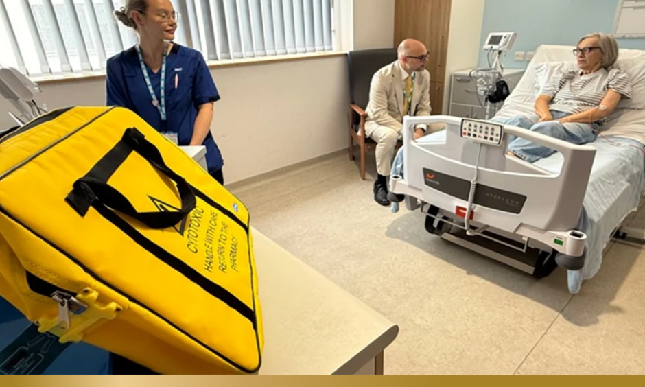 A hospital room with a patient resting in a bed while a healthcare worker and another visitor sit nearby. In the foreground, a bright yellow medical transport bag sits on a table, with monitoring equipment and clinical furnishings visible around the room.