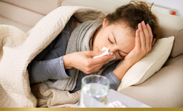 Woman who is sick with COVID-19 lays on a sofa with a blanket sneezing into a tissue