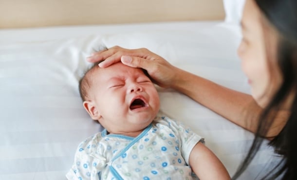 Crying infant with a caregiver placing a hand on their forehead while resting in bed.