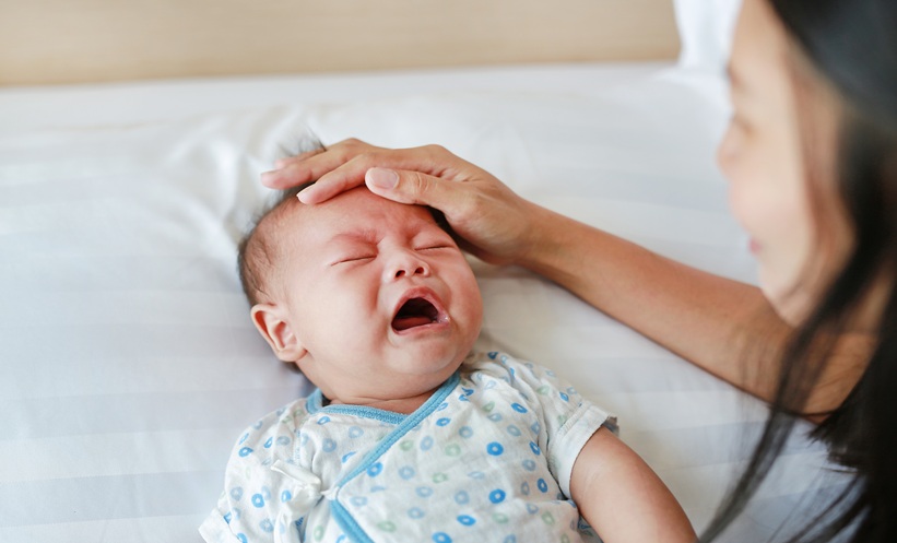 Crying infant with a caregiver placing a hand on their forehead while resting in bed.