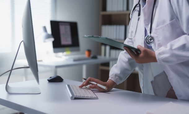 Doctor standing with clipboard and pen looks at results on computer