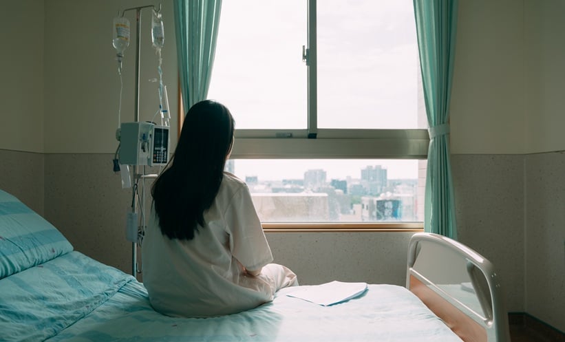 Young woman sits on a hopsital bed and looks out of the window after being diagnosed with cancer